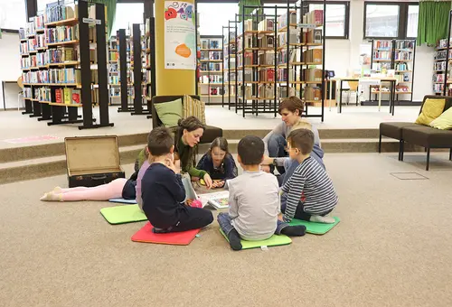 Sechs Kinder sitzen in der Stadtbücherei auf dem Boden und lassen sich etwas von einer Frau vorlesen.