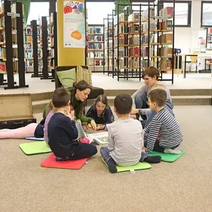 Sechs Kinder sitzen in der Stadtbücherei auf dem Boden und lassen sich etwas von einer Frau vorlesen.