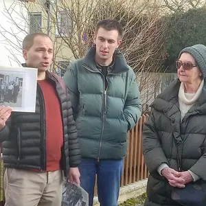 Ein Mann hält eine historische Fotografie hoch, neben ihm stehen ein junger Mann und eine ältere Dame