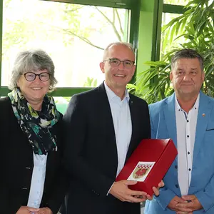 Zwei Männer und eine FRau lächeln in die Kamera. Der mittlere Mann hat ein rotes Präsent erhalten.