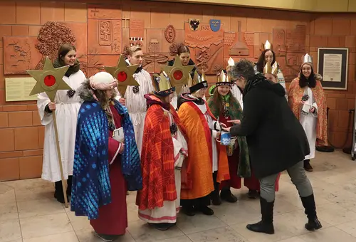 11 Sternsinger in Gewändern erhalten von der Bürgermeisterin eine Spende überreicht.