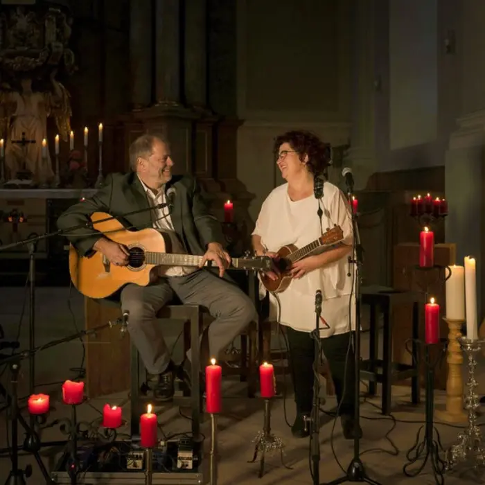 Mann mit Gitarre, Frau mit Ukulele stehen umgeben von Kerzen vor einem Altar und lächeln einander zu.