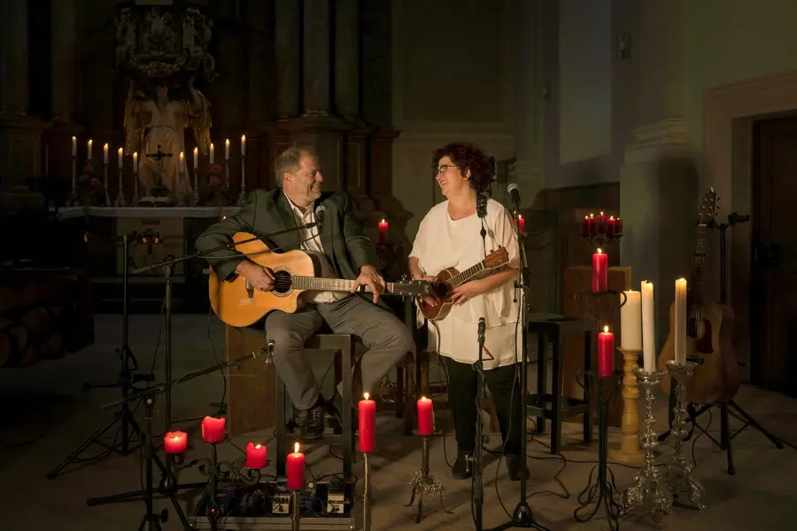 Mann mit Gitarre, Frau mit Ukulele stehen umgeben von Kerzen vor einem Altar und lächeln einander zu.