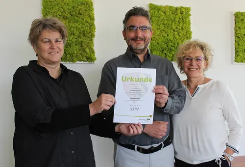 Ein Mann zeigt seine Ernennungsurkunde, zwei Frau rechts und links neben ihm.