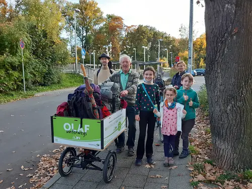 Lastenfahrrad OLAF ist vollgeladen mit Reisegepäck. Im Hintergrund die Familien.