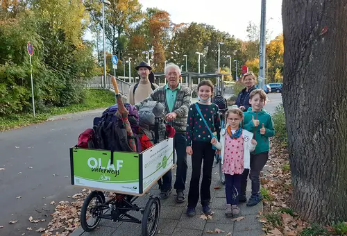 Lastenfahrrad OLAF ist vollgeladen mit Reisegepäck. Im Hintergrund die Familien.