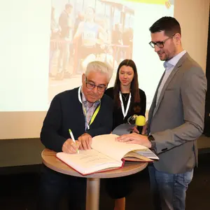 Ein Mann trägt sich in ein Buch ein, während ein anderer Mann daneben steht und eine Frau im Hintergrund zusieht.