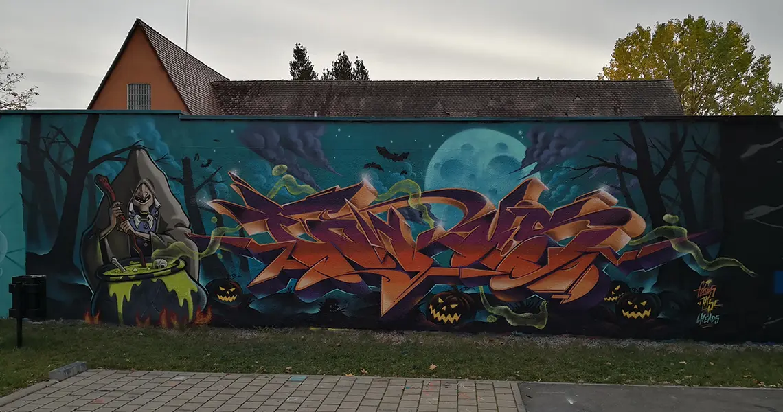 Das Gafittizeigt eine Halloween Szene mit einer Hexe im dunklen Wald mit Bäumen ohne Blätter. Vor ihr steht ein Kessel auf dem Feuer und um sie herum liegen im dnklen Kürbisse mit funkelnden Augen.