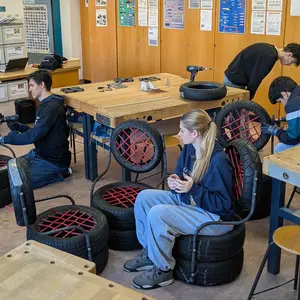 Schüler bearbeiten die Stühle. Eine Schülerin sitzt auf einem der fertigen Stühle. 