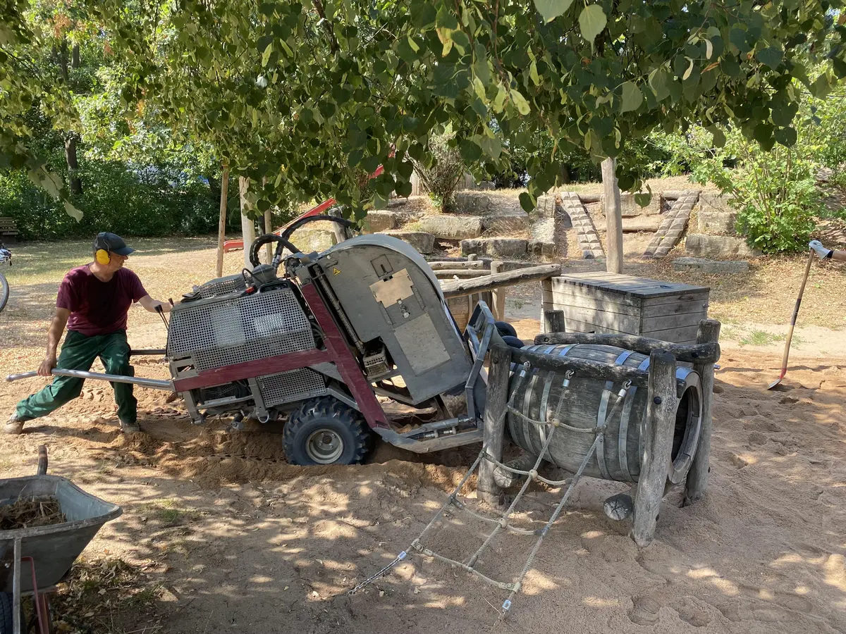 Arbeiter mit Gehörschutz bewegt eine große und schwere Maschine für die Sandreinigung über einen Spielplatz