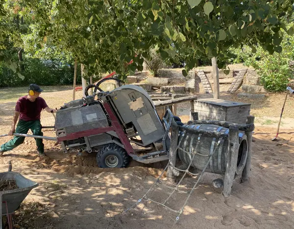Arbeiter mit Gehörschutz bewegt eine große und schwere Maschine für die Sandreinigung über einen Spielplatz