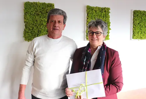 Ein Mann und eine Frau stehen nebeneinander und blicken in die Kamera. Die Frau hält einen Geschenkumschlag.