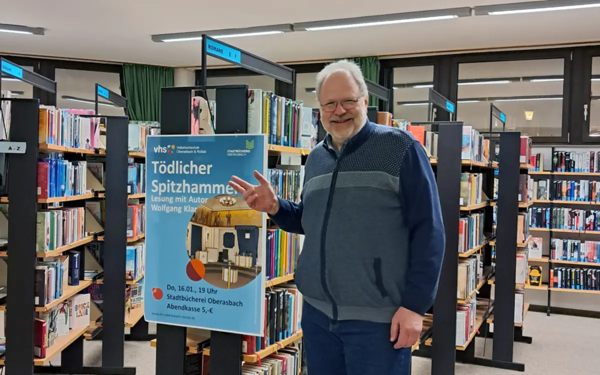 Weißhaariger Mann mit Bart und Brille vor Bücherregalen mit einem Veranstaltungsplakat 
