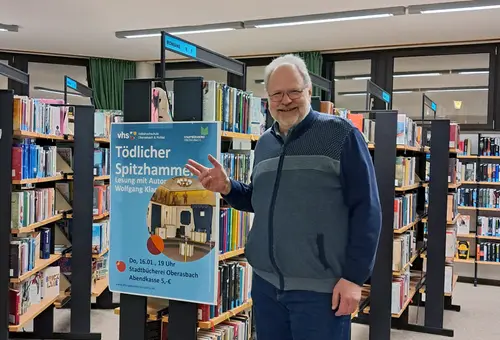 Weißhaariger Mann mit Bart und Brille vor Bücherregalen mit einem Veranstaltungsplakat 