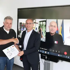 Ein Mann überreicht einenm anderen eine Dankurkunde. Im HIntergrund auf dem Bildschirm sind der Bürgermeister von Solotschiw und der Übersetzer zu sehen.