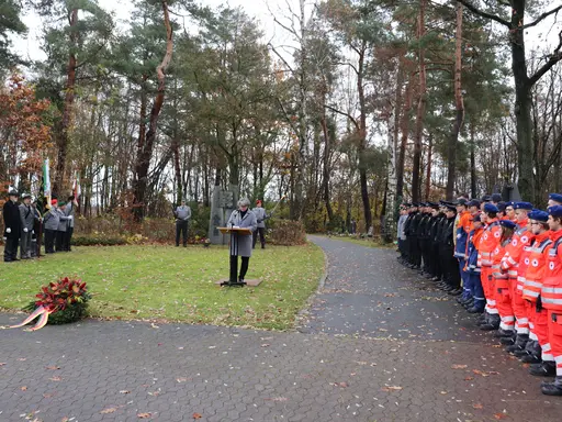 Birgit Huber hält eine Rede auf dem Friedhof. Links und rechts Abordnungen, im Vordergrund ein Kranz.