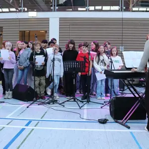 Kinder singen im Chor; davor eine Lehrerin am Keyboard