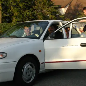 Weißes Auto mit mehreren Personen, eine weitere Frau steigt zu.