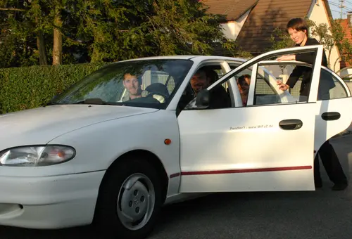 Weißes Auto mit mehreren Personen, eine weitere Frau steigt zu.
