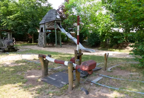 Schienen mit einer Schrankenanlage auf einem Spielplatz unter Bäumen.