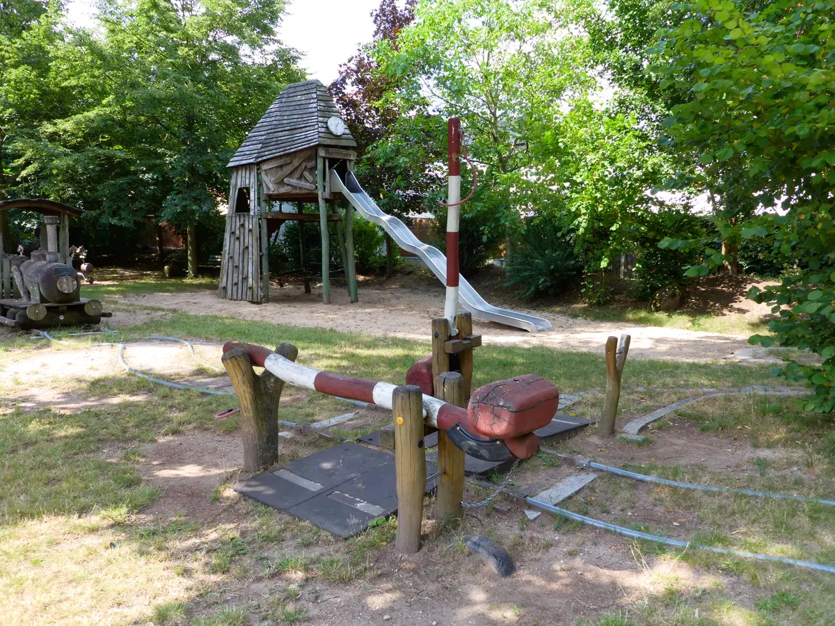 Schienen mit einer Schrankenanlage auf einem Spielplatz unter Bäumen.