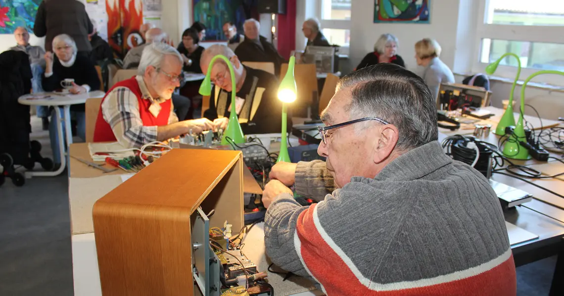 Repair Café, ein Mann repariert ein altes Radio, weitere Personen im Hintergrund bei Kaffee und Kuchen im Gespräch