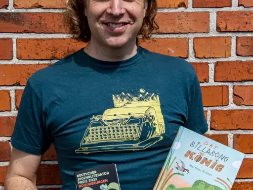 Matthias Kröner vor Backsteinmauer. Er hält sein Buch Der Billabongkönig in der linken Hand und in der rechten eine Broschüre zur Nominierung für den Kinder- und Jugendbuchpreis. 