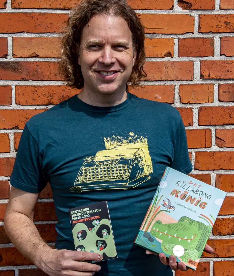 Matthias Kröner vor Backsteinmauer. Er hält sein Buch Der Billabongkönig in der linken Hand und in der rechten eine Broschüre zur Nominierung für den Kinder- und Jugendbuchpreis. 