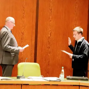 Zwei Männer vor einer Holzwand. Einer hebt die Hand zum Schwur. 