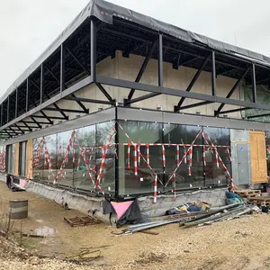Blick auf die Baustelle der Dreiachsporthalle. Gebäude und Dach sind komplett, die Fassade ist verglast. Davor noch rot-weiße Flatterbänder zur Vogelabwehr. 
