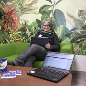 Birgit Huber sitzt mit Tablet auf einem grünen Sofa vor einer Dschungeltapete. Vor ihr auf dem Sofatisch steht ein Laptop auf dessen Monitor das Bürgerserviceportal zu sehen ist. 