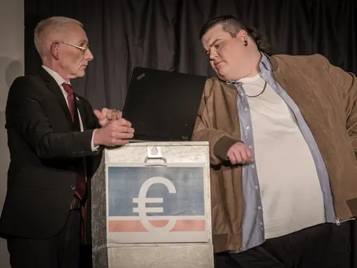 Zwei Männer schauen auf Laptop. Einer im Anzug, einer in Lederjacke mit Goldkette.