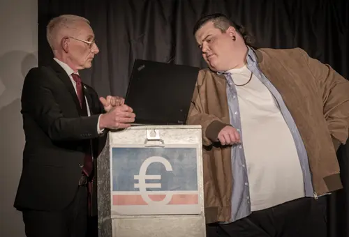 Zwei Männer schauen auf Laptop. Einer im Anzug, einer in Lederjacke mit Goldkette.