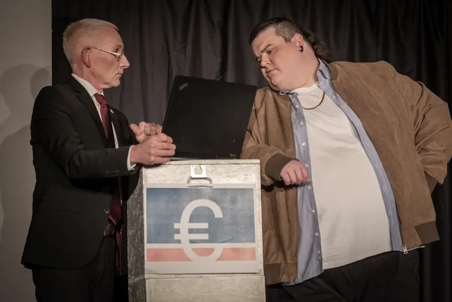 Zwei Männer schauen auf Laptop. Einer im Anzug, einer in Lederjacke mit Goldkette. 