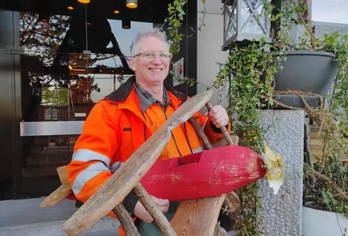 Ein Mann in orangener Arbeitskleidung hält ein kleines, selbstgebautes Doppeldecker-Flugzeug.