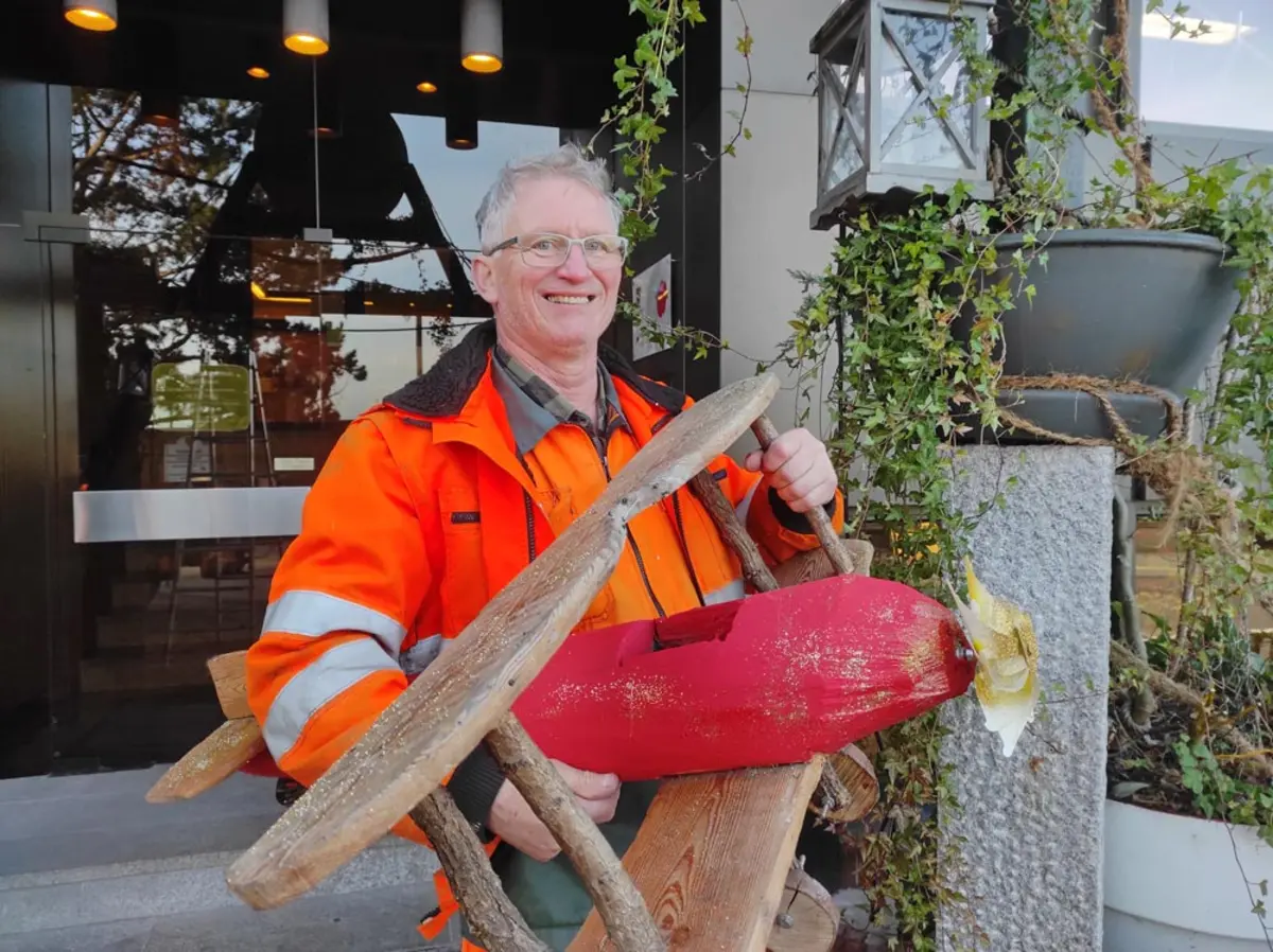 Ein Mann in orangener Arbeitskleidung hält ein kleines, selbstgebautes Doppeldecker-Flugzeug.