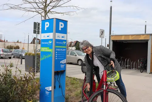 Frau mit E-Bike an der blauen E-Ladesäule für E-Bikes.