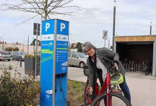 Frau mit E-Bike an der blauen E-Ladesäule für E-Bikes.
