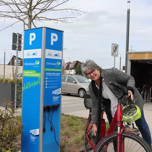 Frau mit E-Bike an der blauen E-Ladesäule für E-Bikes.