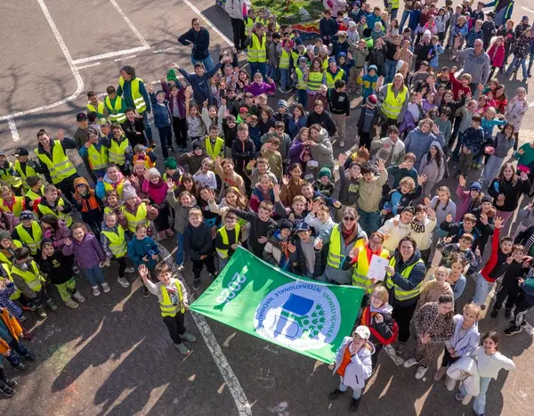 Eine große Menge an Grundschülern mit Eltern und Lehrern, viele in gelben Warnwesten, die eine ausgerollte Fahne halten
