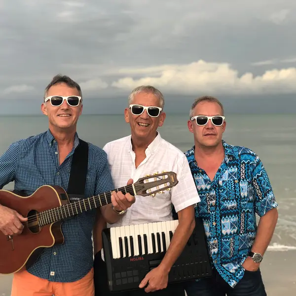 Band aus drei Männern mit Gitarre in der Hand und Keyboard-Tastatur unterm Arm am Strand. Alle tragen Sonnenbrillen und lächeln in die Kamera. 