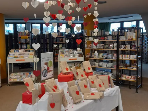 Tisch mit einem weißen Tuch geschmückt. Darauf die Bücher fürs Blind Date in braunen Tüten. Über dem Tisch hängen Girlanden aus roten und weißen Herzen. 