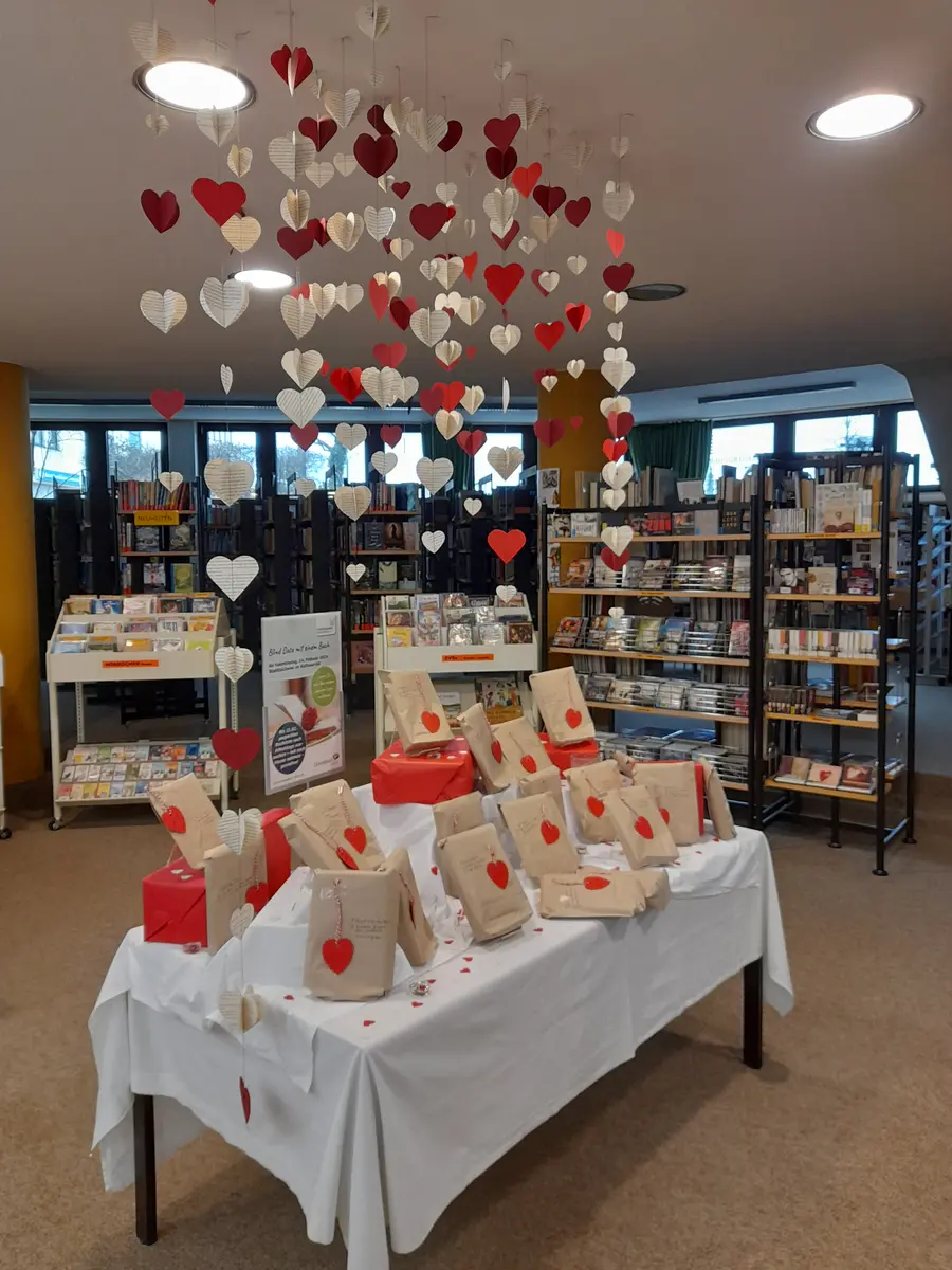 Tisch mit einem weißen Tuch geschmückt. Darauf die Bücher fürs Blind Date in braunen Tüten. Über dem Tisch hängen Girlanden aus roten und weißen Herzen.