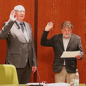 Zwei Männer vor einer Holzwand. Beide heben die rechte Hand zum Schwur. 
