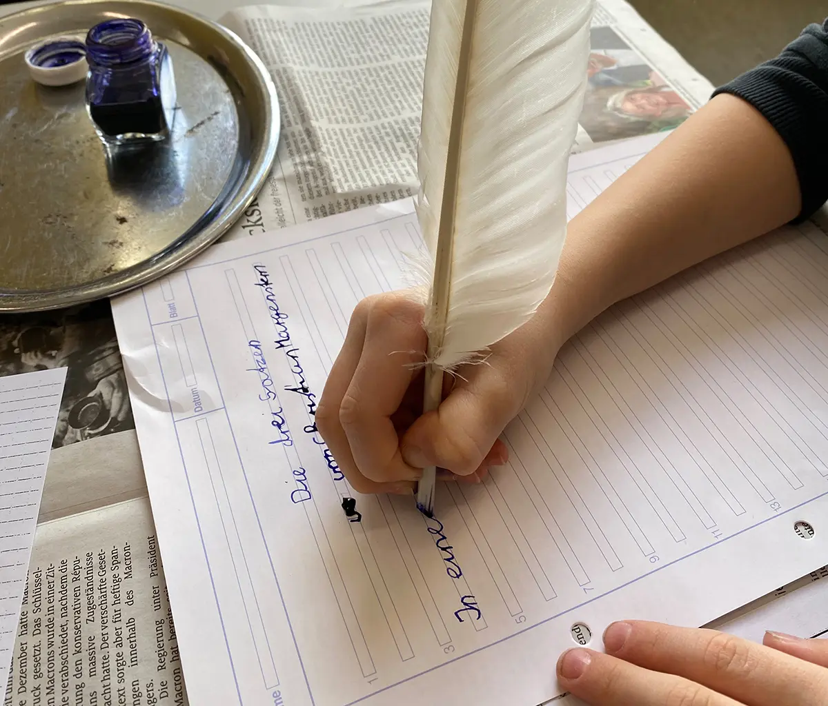 Eine Kinderhand schreibt auf einem Blatt Papier mit einer Gänsefeder und Tinte.