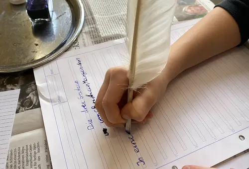 Eine Kinderhand schreibt auf einem Blatt Papier mit einer Gänsefeder und Tinte.