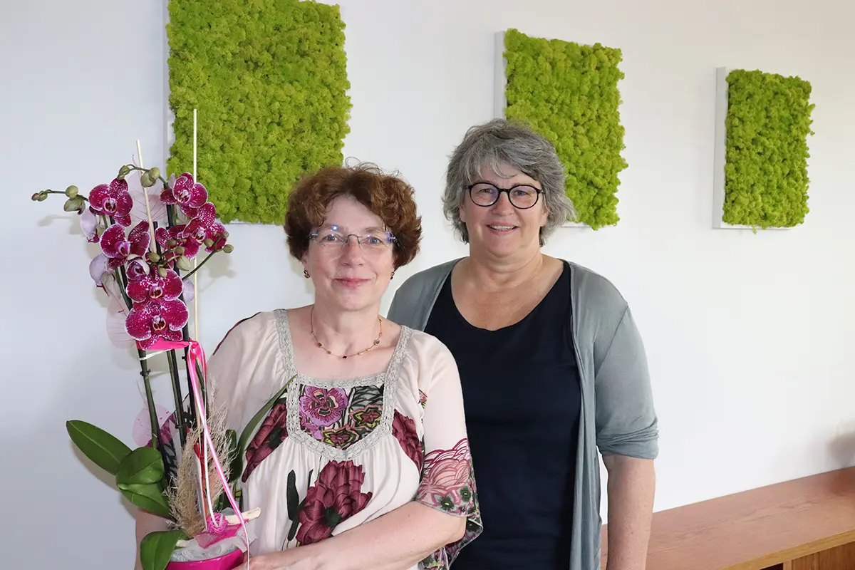 Zwei Frauen vor einer weißen Wand. Eine FRau trägt eine Orchidee.