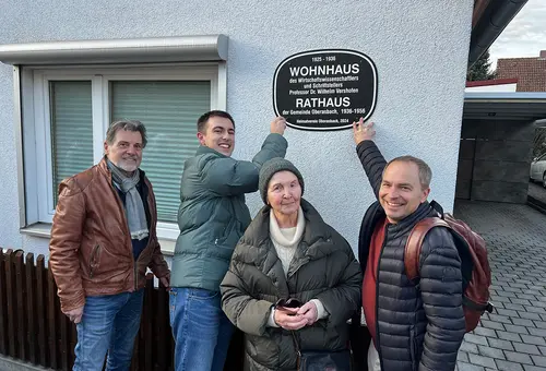 Zwei Männer halten ein Schild an eine Hauswand und befestigen es mit Schrauben. Ein Mann und eine Frau schauen zu.