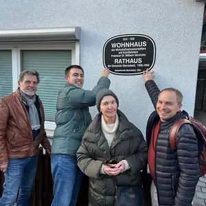 Zwei Männer halten ein Schild an eine Hauswand und befestigen es mit Schrauben. Ein Mann und eine Frau schauen zu.