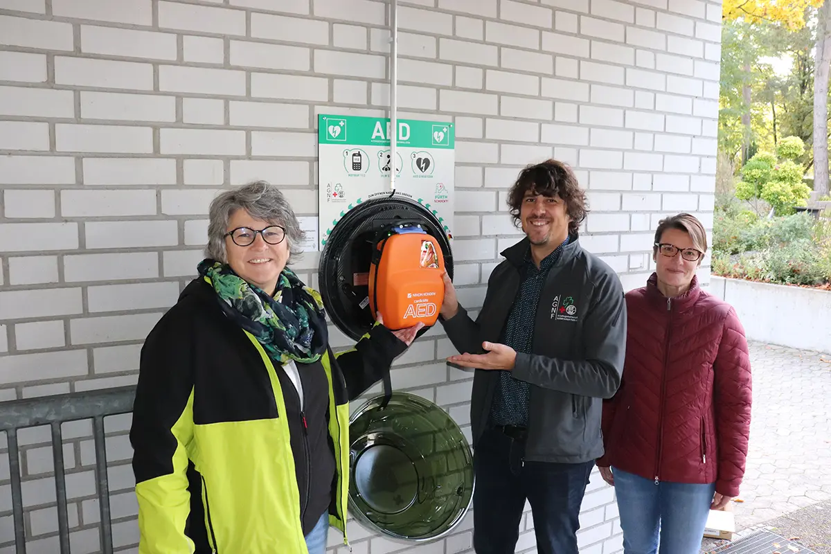 Zwei Frauen und ein Mann zeigen auf den neuen Wand-Defibrillator.